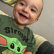 Kaizen is registered to the contest to win money with this photo: baby_yoda, background, child, clothing, couch, cute, eyes, face, green_tshirt, happy, indoor, painting, person, portrait, short_hair, sitting, smiling, teeth, toddler, wall