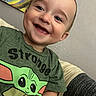 baby_yoda, background, child, clothing, couch, cute, eyes, face, green_tshirt, happy, indoor, painting, person, portrait, short_hair, sitting, smiling, teeth, toddler, wall