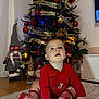 baby, christmas_tree, festive_clothing, red_socks, holiday_decorations, gnome_figurines, carpet, indoor, christmas, ornaments, pine_cones, ribbon, wooden_floor, child, smiling, cute, holiday, sitting, toddler, toy