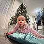 baby, infant, christmas_tree, holiday_decorations, pink_clothing, blue_mat, floor, home_interior, curtains, table, chair, plant, window, lights, ornaments, star, candy_cane, heart_ornament, person, cute