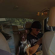 Draco joined the competition — help win amazing prizes! dog, person, car_interior, seat, window, trees, sunlight, casual_clothing, sunglasses, headwear, resting, leather_seat, seatbelt, pet, companion, daytime, relaxed, technology, phone, portrait