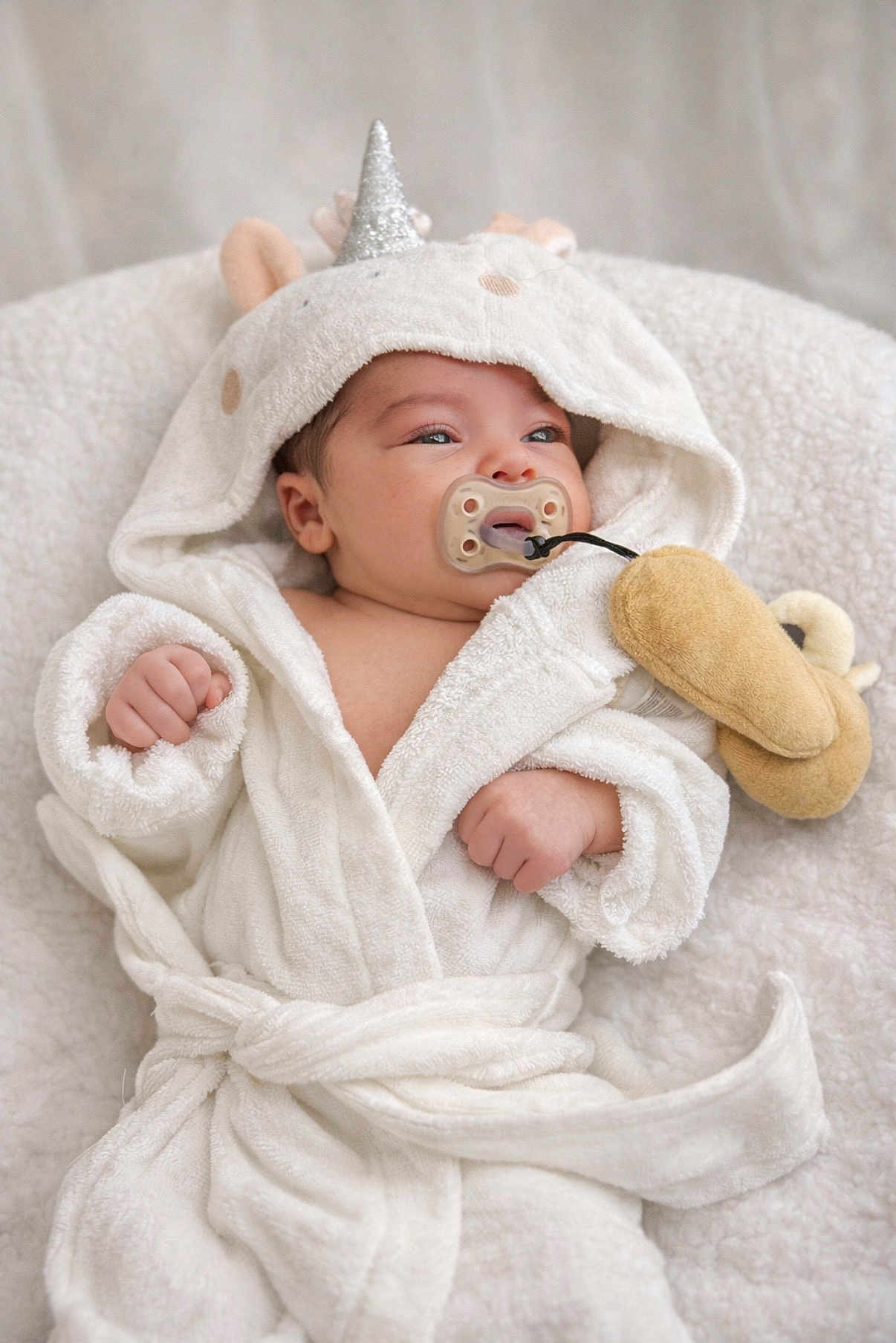 Kiara is registered to the contest to win money with this photo: baby, infant, newborn, pacifier, hood, unicorn, bathrobe, robe, blanket, plush_toy, soft_texture, cheeks, hand, portrait, indoors, cute, sleepy, wrapped, eyelids, towel