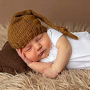 Jordan joined the competition — help win amazing prizes! baby, newborn, sleeping, hat, knitted_hat, pillow, white_onesie, fluffy_rug, brown, cozy, infant, resting, peaceful, cute, soft_texture, portrait, child, indoors, closeup, sleep