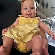 Aylani joined the competition — help win amazing prizes! baby, child, yellow_dress, tongue_out, smiling, person, indoor, living_room, furniture, couch, blanket, hand, skin, cute, happy, infant, portrait, seated, adult_hands, cozy