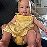 baby, child, yellow_dress, tongue_out, smiling, person, indoor, living_room, furniture, couch, blanket, hand, skin, cute, happy, infant, portrait, seated, adult_hands, cozy