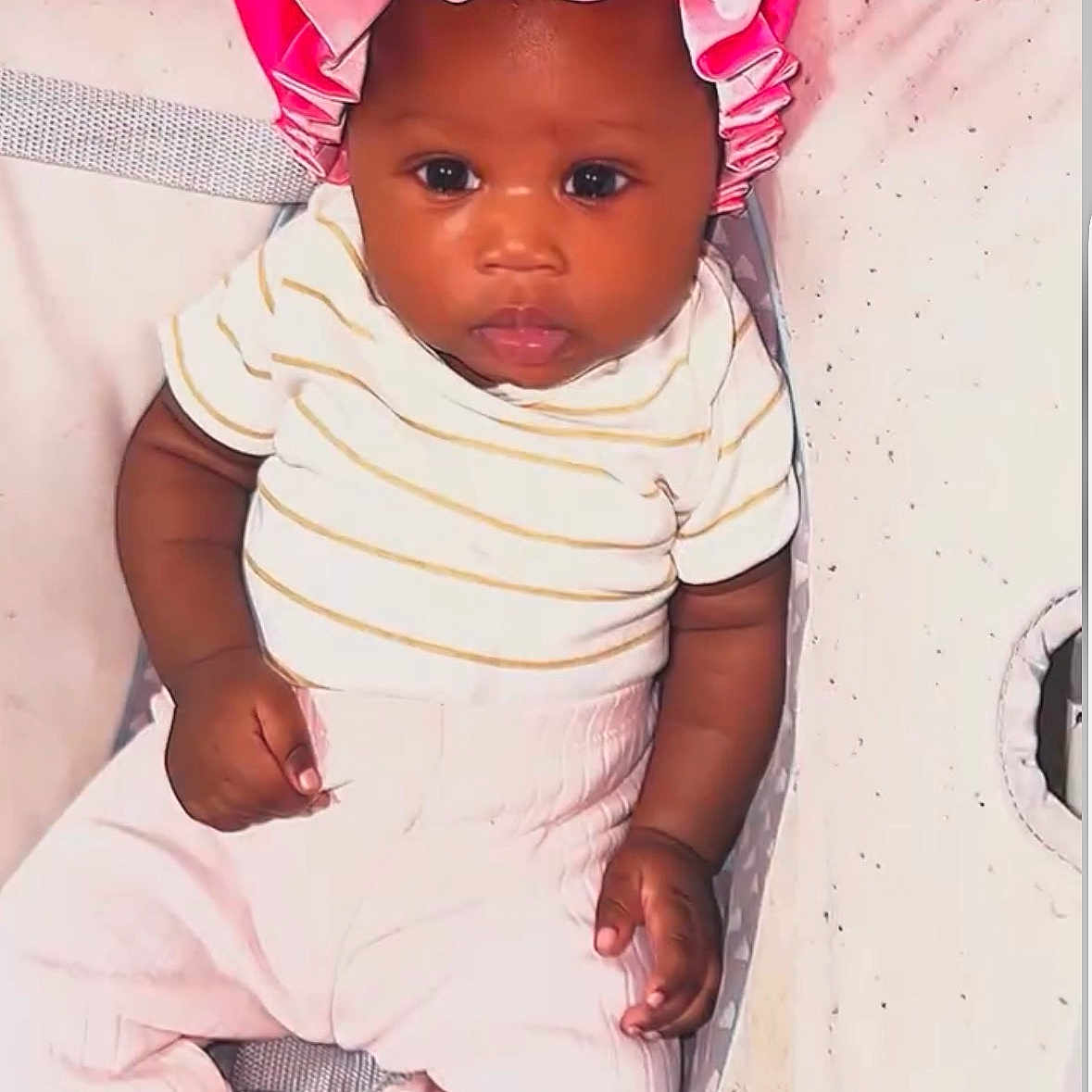 Jaziriah is registered to the contest to win money with this photo: baby, infant, pink_bonnet, striped_shirt, pants, cushion, face, child, person, indoor, clothing, cute, young, portrait, seated, looking_at_camera, skin, headwear, baby_girl, comfort