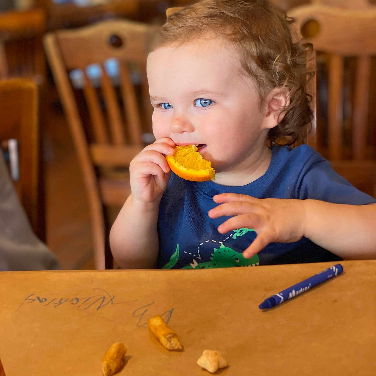 Nicklas joined the competition — help win amazing prizes! background_blur, blue_eyes, brown_paper, casual_clothing, chair, child, crayon, curly_hair, cute, eating, food, happy, indoor, orange, person, portrait, snack, table, toddler, wooden_furniture