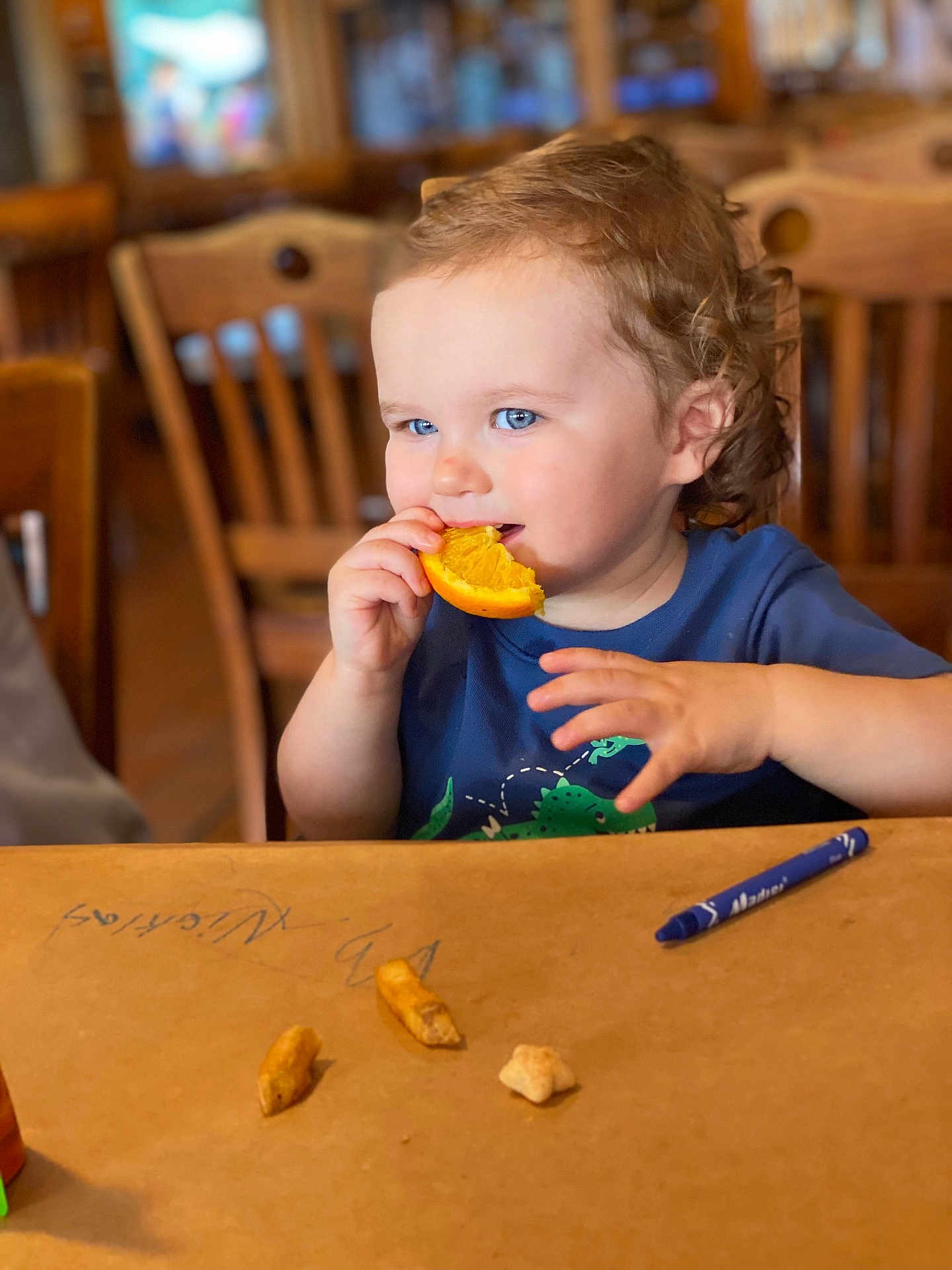 Nicklas joined the competition — help win amazing prizes! toddler, child, orange, food, snack, curly_hair, blue_eyes, table, brown_paper, crayon, chair, wooden_furniture, indoor, eating, cute, person, portrait, happy, casual_clothing, background_blur