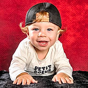 Maëlan participe au concours pour gagner de l'argent avec cette photo : toddler, child, smiling, blue_eyes, curly_hair, cap, black_cap, white_shirt, text_on_shirt, hands, lying_down, red_background, fur_surface, portrait, baby, cute, happy, indoor, close_up, face