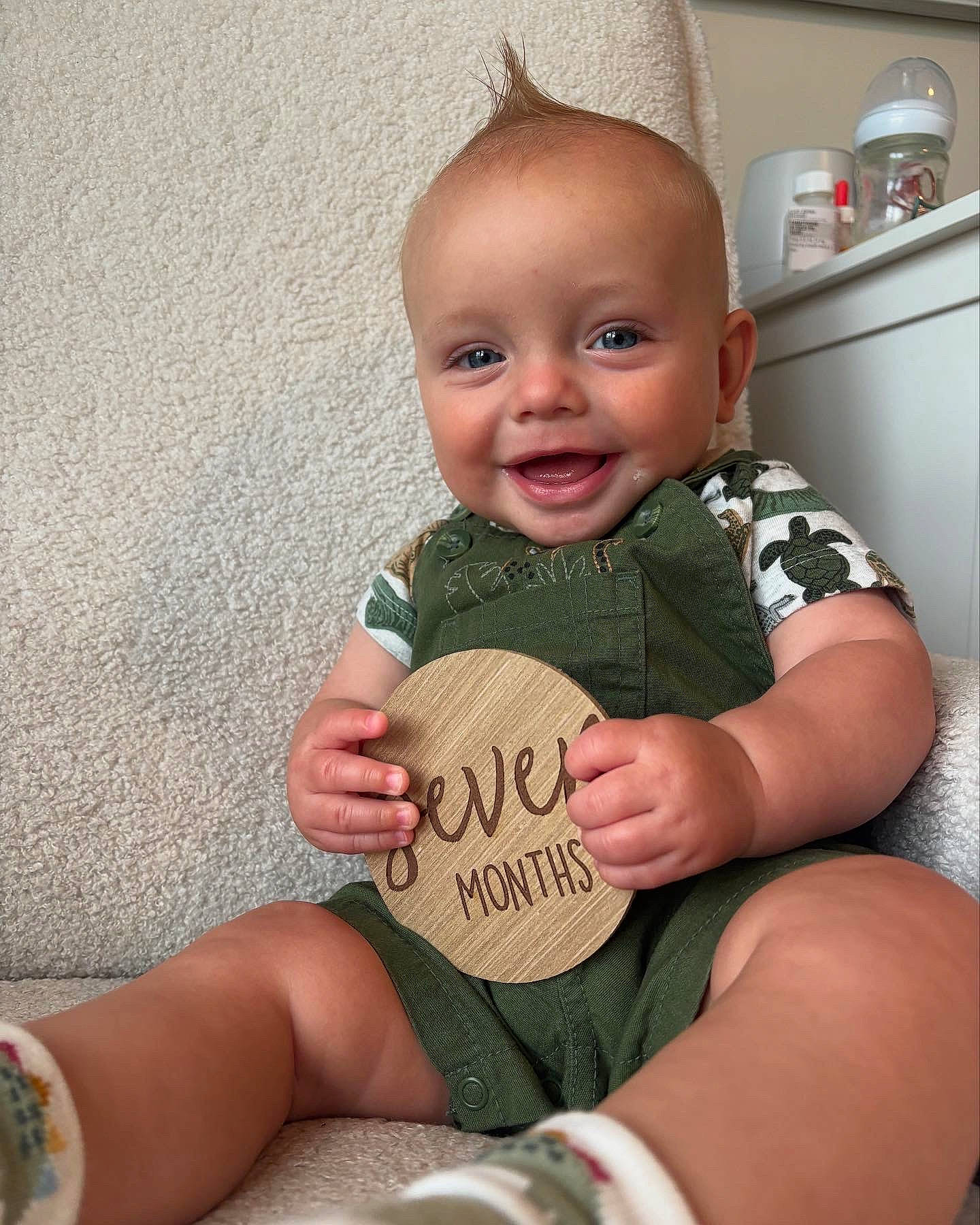 Luca joined the competition — help win amazing prizes! baby, baby_clothes, child, cute, expression, face, feet, green_overalls, hand, happy, indoor, infant, milestone, person, plush_chair, portrait, sitting, skin, smiling, wooden_plaque