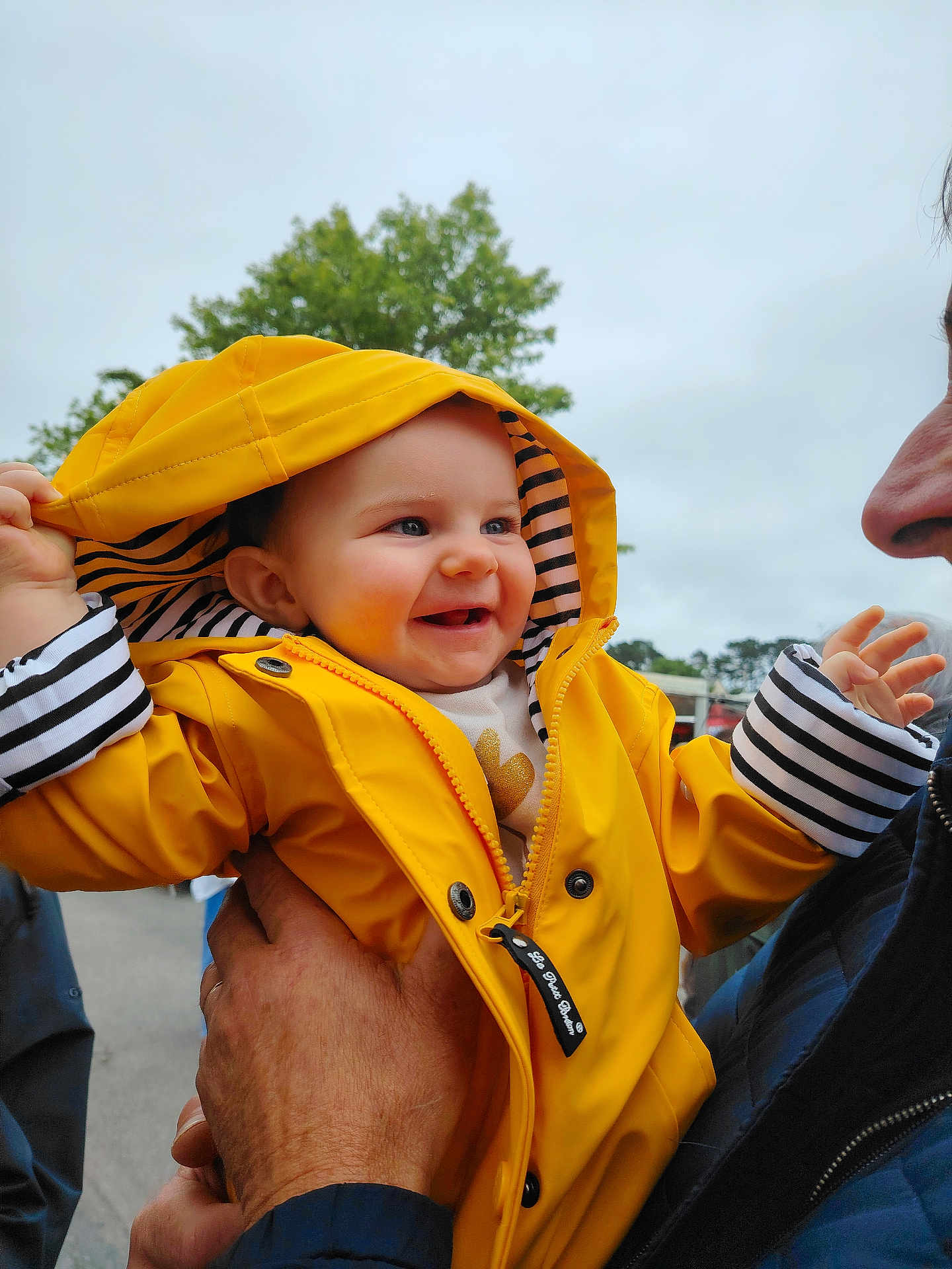 Margot a rejoint le concours — aidez-le/la à gagner de superbes lots ! baby, child, smiling, yellow_raincoat, hood, stripes, outdoor, tree, adult_hand, happy, face, person, jacket, cloudy_sky, holding, cute, portrait, clothing, expression, nature