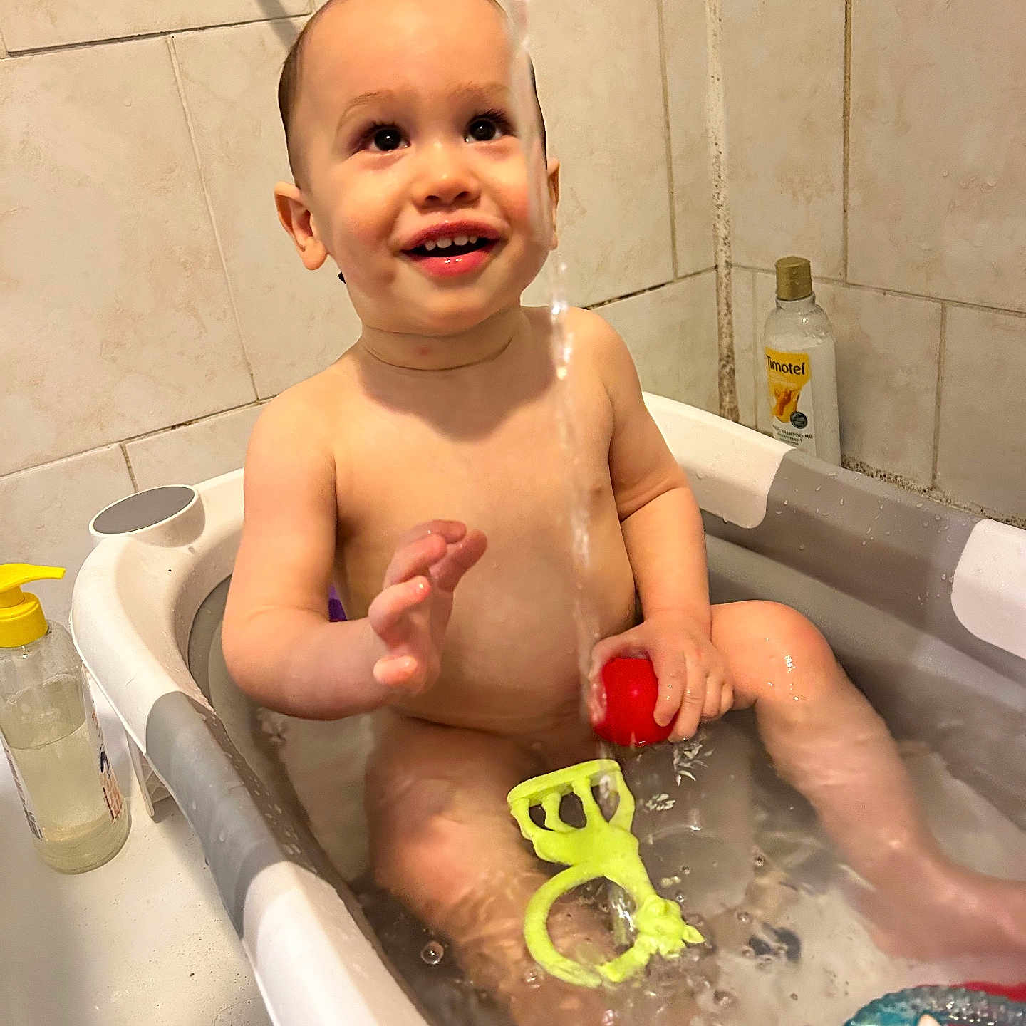 Tilio participe au concours pour gagner de l'argent avec cette photo : baby, bath, bathroom, bathtub, child, green_teether, happy, playing, red_ball, shampoo_bottle, shower_head, sitting, smile, soap_dispenser, splash, tiled_wall, toddler, toy, undressed, water