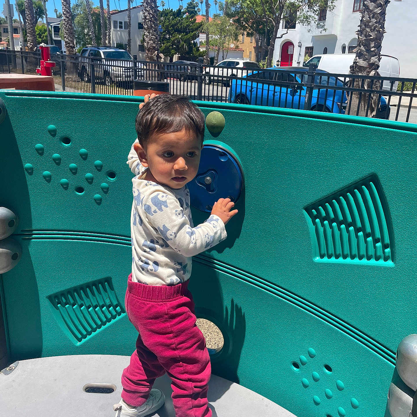 Elliot is registered to the contest to win money with this photo: car, casual, child, clothing, curious, daytime, fence, outdoor, palm_trees, park, person, play_equipment, playground, sneakers, standing, sunlight, toddler, toy, tree, urban