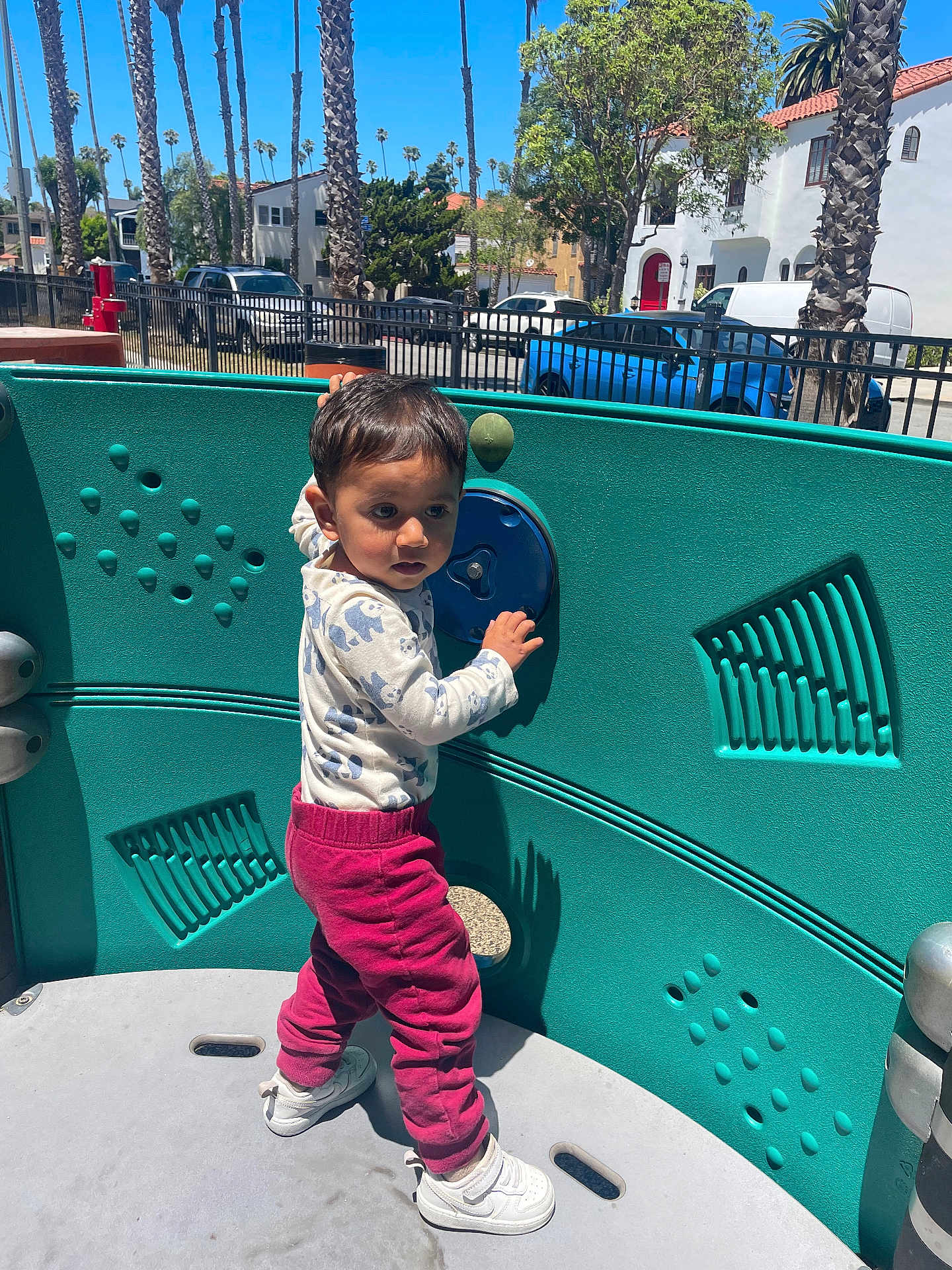 Elliot is registered to the contest to win money with this photo: toddler, child, playground, outdoor, sunlight, clothing, sneakers, palm_trees, toy, play_equipment, urban, park, fence, car, tree, person, daytime, curious, standing, casual