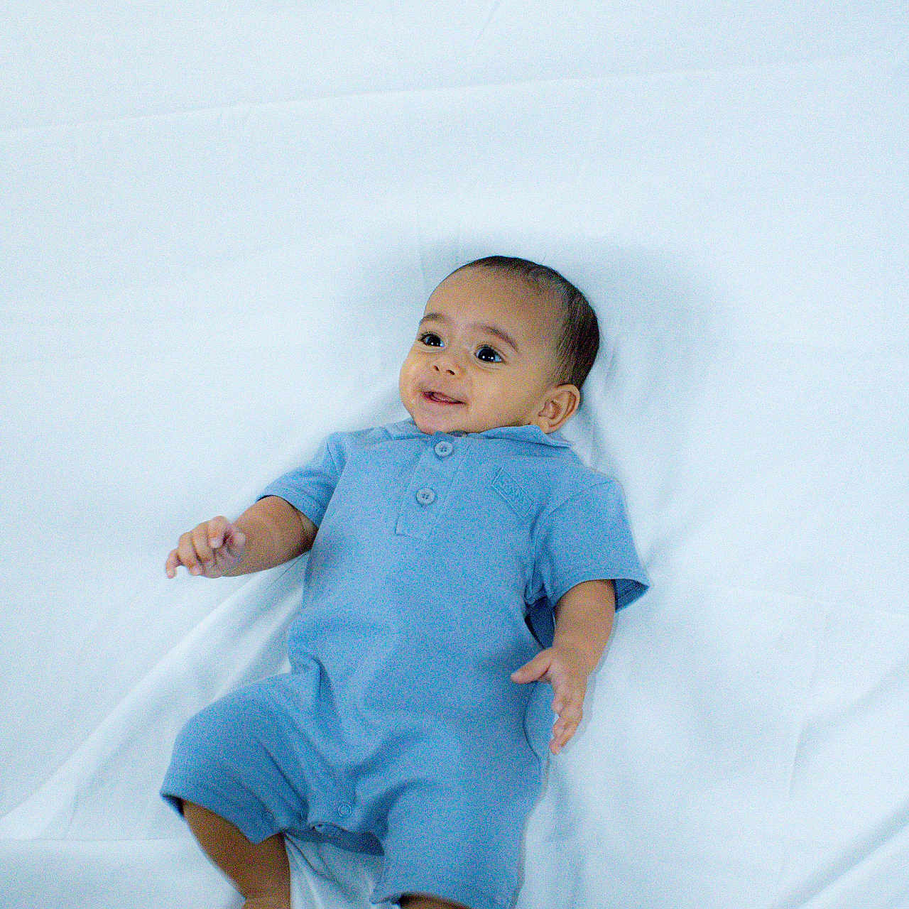 Asaiah is registered to the contest to win money with this photo: baby, infant, child, portrait, lying_down, blue_outfit, romper, blanket, white_background, bed, smiling, happy, face, eyes, hands, feet, indoor, photography, candid, baby_portrait
