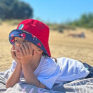Mylann a rejoint le concours — aidez-le/la à gagner de superbes lots ! beach, blue_sky, casual_clothing, child, daylight, hat, outdoor, person, portrait, red_hat, relaxing, resting, sand, sky, summer, thinking, towel, vacation, white_shirt, young