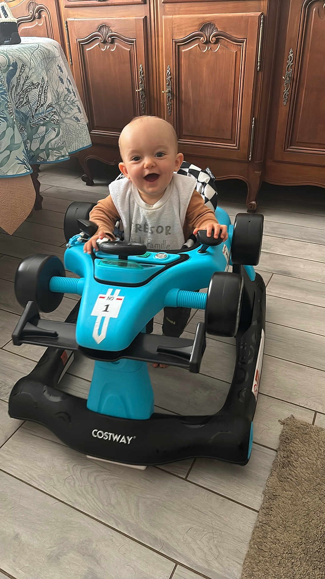 Nino a rejoint le concours — aidez-le/la à gagner de superbes lots ! baby, child, toy_car, blue, indoor, floor, wooden_furniture, smiling, steering_wheel, tiled_floor, play, happy, person, clothing, cabinet, car_seat, car_toy, infant, floor_rug, hands