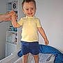 child, toddler, smiling, standing, yellow_shirt, blue_shorts, barefoot, changing_table, hand_holding, nursery, diapers, shelf, baby_supplies, indoor, person, happy, hair_standing_up, light_skin, blue_eyes, casual_clothing