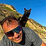 man, sunglasses, black_cat, cat, pet, beach, cliff, blue_sky, seagulls, hillside, sunny, smile, mustache, portrait, harness, outdoors, vacation, sand, reflection, close_up