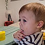 Henni a rejoint le concours — aidez-le/la à gagner de superbes lots ! toddler, child, indoor, striped_shirt, table, yellow_container, curious, face, hand, fingers, lamp, clock, artwork, wall, hair, person, seated, room, furniture, portrait