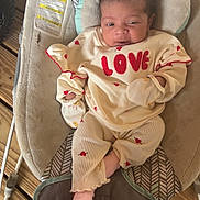 Lucia is registered to the contest to win money with this photo: baby, infant, newborn, onesie, love_text, face, head, eyes, smile, mittens, sock, baby_bouncer, cushion, seat, wooden_floor, patterned_fabric, clothing, cozy, indoor, portrait