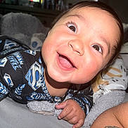Bryce is registered to the contest to win money with this photo: baby, smiling, superman, onesie, child, happy, portrait, indoors, blanket, stuffed_animal, face, cute, infant, closeup, person, lying_down, sleepwear, bright_eyes, skin, hand