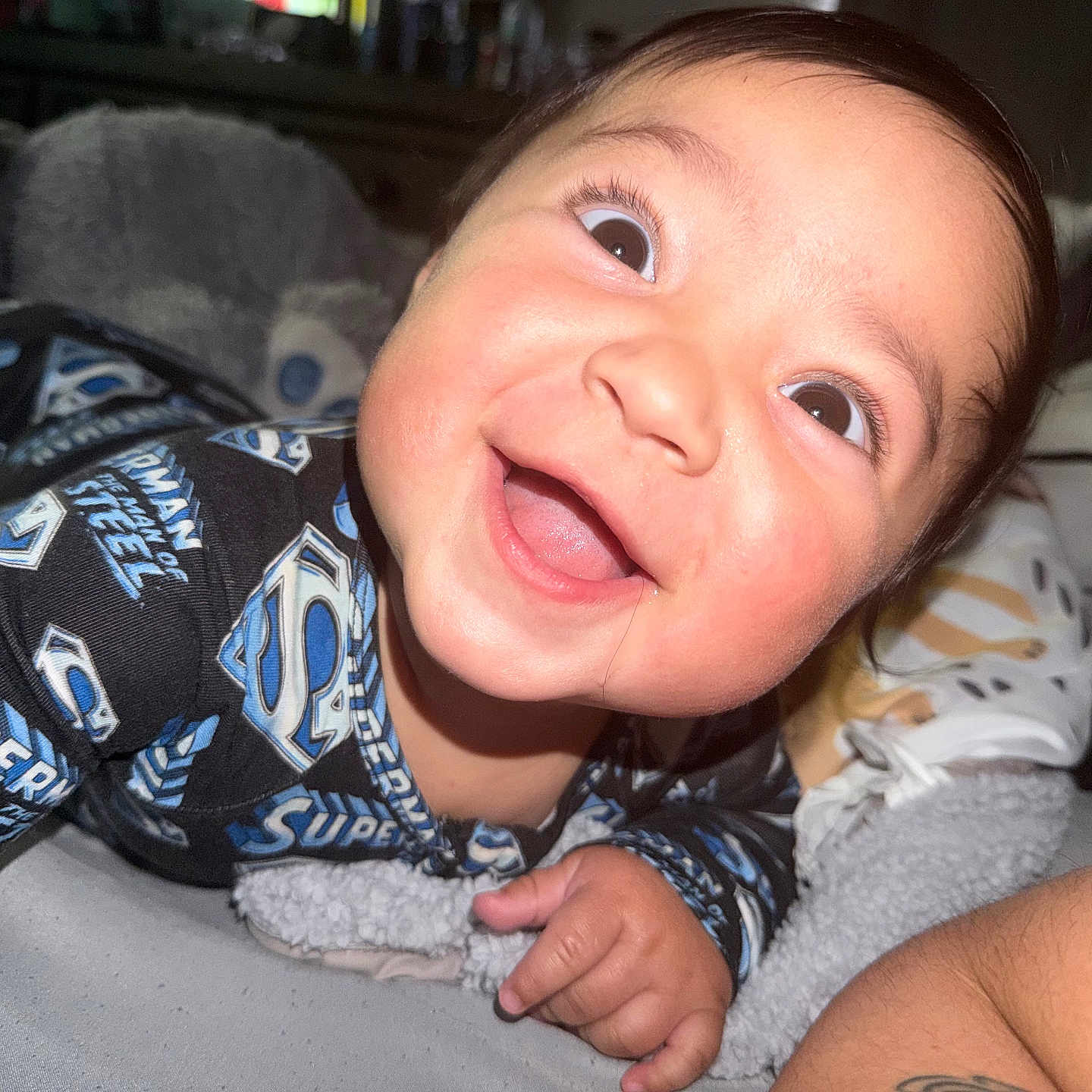 Bryce is registered to the contest to win money with this photo: baby, smiling, superman, onesie, child, happy, portrait, indoors, blanket, stuffed_animal, face, cute, infant, closeup, person, lying_down, sleepwear, bright_eyes, skin, hand