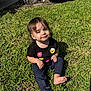 toddler, child, grass, outdoor, sunlight, black_shirt, smiley_face, flower_design, barefoot, casual_clothing, greenery, nature, young_child, sitting, daylight, cute, happy, portrait, person, playful