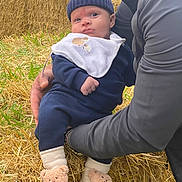 Elliot joined the competition — help win amazing prizes! baby, infant, child, hat, knit_hat, bib, socks, slippers, teddy_bear, person, adult_hand, straw, hay_bale, grass, outdoor, casual_clothing, blue_clothing, cute, warm_clothing, holding