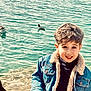 child, boy, smile, curly_hair, denim_jacket, water, lake, ducks, outdoor, nature, casual_clothing, happy, portrait, young, people, animal, bird, sunlight, face, shore