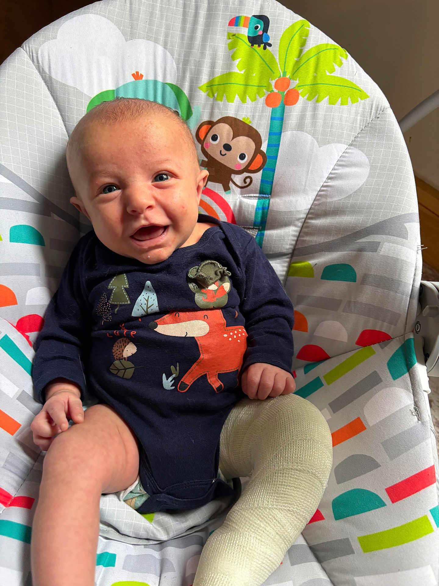 Reese joined the competition — help win amazing prizes! baby, infant, cast, leg_cast, smiling, blue_onesie, animal_print, baby_chair, monkey, toucan, palm_tree, colorful, indoor, happy, cute, child, sitting, clothing, patterned, playful