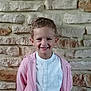 child, smiling, boy, pink_hoodie, white_shirt, short_hair, curly_hair, stone_wall, background, casual_clothing, standing, portrait, happy, indoors, young, cute, person, fashion, buttoned_shirt, light
