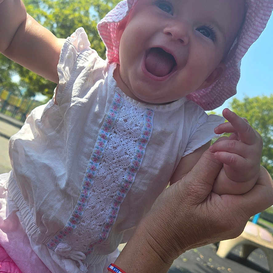 Violet is registered to the contest to win money with this photo: arm, baby, blue_sky, bracelets, child, daylight, face, greenery, happy, holding_hands, joy, outdoor, park, person, pink, playground, skin, smiling, sunhat, white_dress