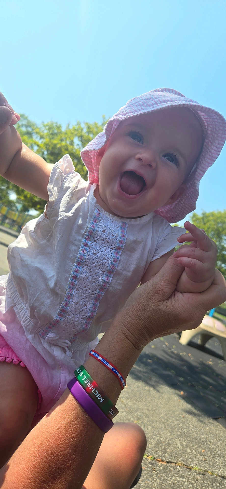 Violet is registered to the contest to win money with this photo: baby, child, smiling, happy, sunhat, white_dress, outdoor, daylight, greenery, person, holding_hands, playground, park, skin, arm, bracelets, pink, blue_sky, face, joy