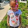 child, girl, braids, beads, tshirt, mickey_mouse, minnie_mouse, outdoor, sidewalk, greenery, plants, sunlight, parking_lot, casual_clothing, smile, standing, daytime, young_child, cute, portrait