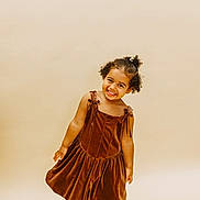 Tiani is registered to the contest to win money with this photo: child, girl, smiling, brown_dress, curly_hair, black_shoes, standing, studio, portrait, happy, playful, fashion, cute, young, one_person, full_body, indoor, light_background, casual, pretty