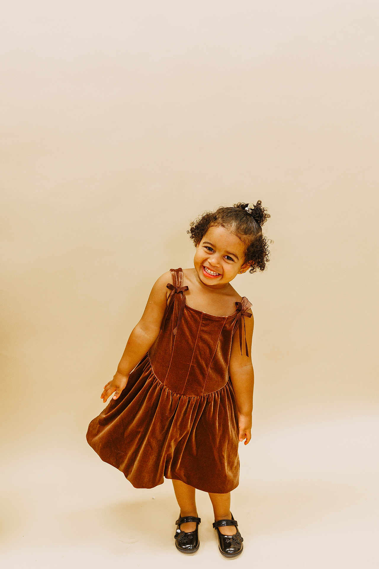 Tiani is registered to the contest to win money with this photo: child, girl, smiling, brown_dress, curly_hair, black_shoes, standing, studio, portrait, happy, playful, fashion, cute, young, one_person, full_body, indoor, light_background, casual, pretty