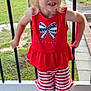child, blonde_hair, red_clothing, striped_shorts, outdoor, smiling, porch, railing, grass, trees, daylight, shoes, socks, cute, happy, person, young_child, casual, summer, playful