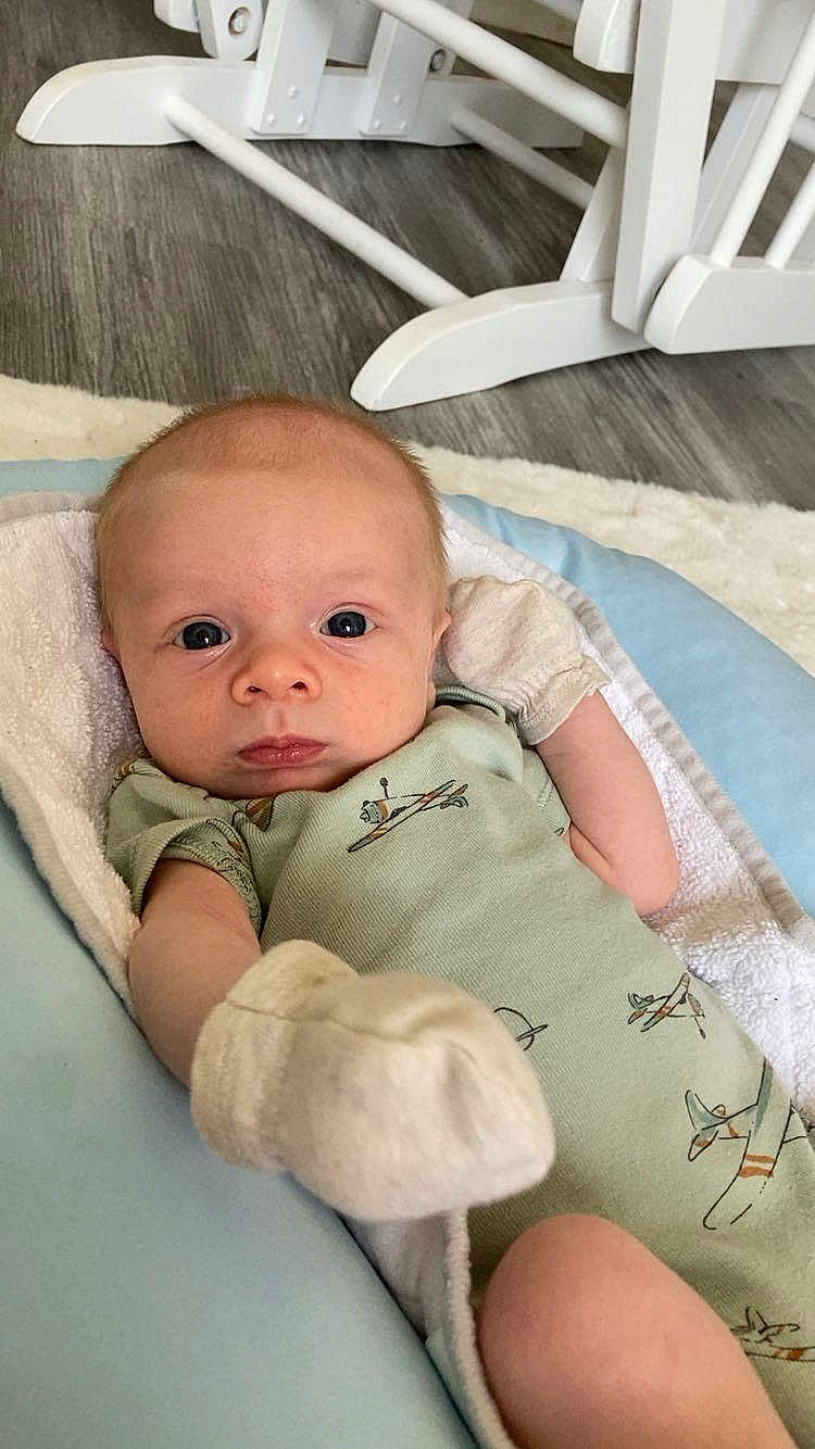 Zachary joined the competition — help win amazing prizes! baby, infant, onesie, mittens, cushion, face, eyes, floor, wooden_chair, rocking_chair, blanket, clothing, person, portrait, indoor, baby_bed, newborn, soft_toy, baby_garment, baby_accessory