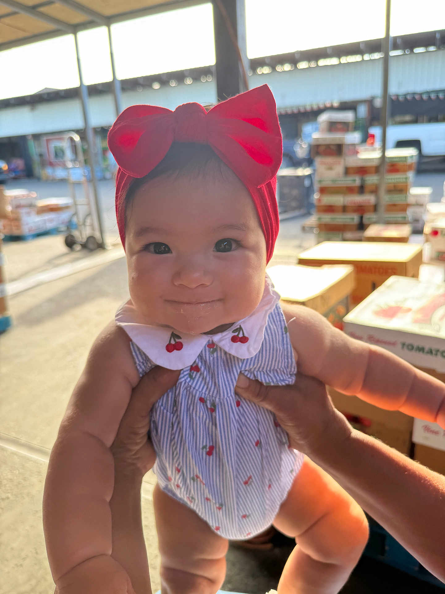 Yareli is registered to the contest to win money with this photo: baby, child, red_bow, headband, smile, drool, hands, romper, cherry_pattern, warehouse, boxes, sunlight, cute, person, infant, holding, outdoor, bright, happy, portrait