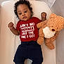 baby, infant, teddy_bear, crib, red_shirt, pants, socks, curly_hair, lying_down, soft_toy, child, cute, comfortable, indoors, portrait, person, smiling, baby_clothing, resting, mesh_sides