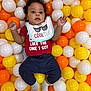 baby, bib, child, colorful_balls, orange, yellow, white, socks, pants, red_shirt, curly_hair, play_area, indoor, cute, portrait, smiling, lying_down, person, fun, soft_lighting