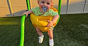 Anthony is registered to the contest to win money with this photo: toddler, swing, child, pacifier, green_swing, yellow_swing, grass, fence, trees, sunlight, outdoor, shoes, legs, happy, smile, playing, park, daylight, person, casual_clothing
