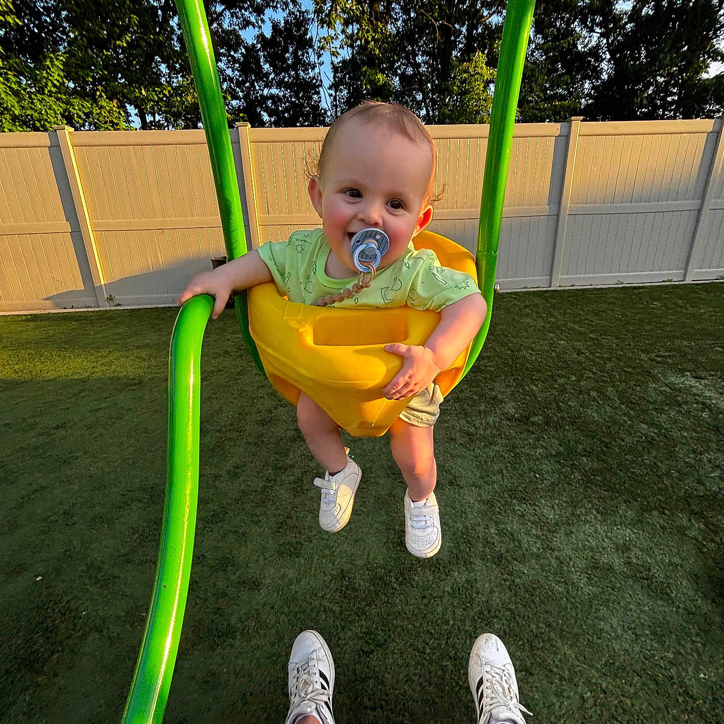 Anthony is registered to the contest to win money with this photo: toddler, swing, child, pacifier, green_swing, yellow_swing, grass, fence, trees, sunlight, outdoor, shoes, legs, happy, smile, playing, park, daylight, person, casual_clothing