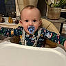 baby, pacifier, high_chair, pajamas, holiday_theme, indoor, plant, potted_plant, window, table, child, face, seated, curious, infant, furniture, home, decor, evening, cozy