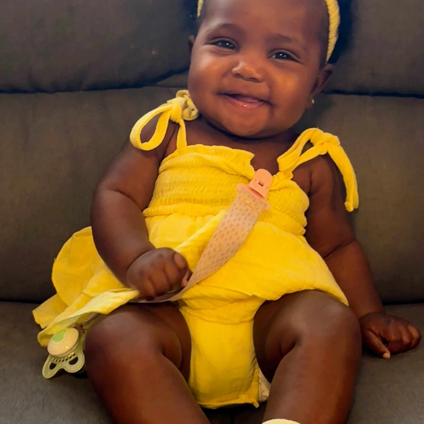 Harmony is registered to the contest to win money with this photo: baby, child, smiling, yellow_dress, headband, pacifier, socks, sitting, couch, indoor, cute, happy, infant, portrait, person, face, skin, clothing, accessory, comfort