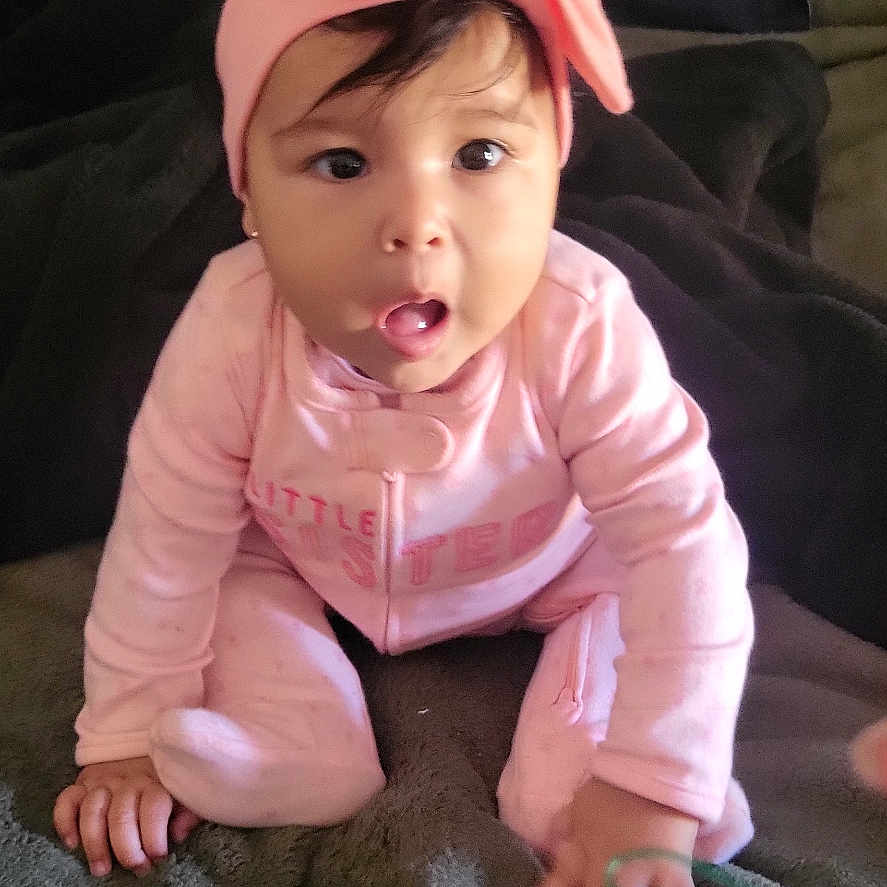 Iris is registered to the contest to win money with this photo: baby, baby_girl, blanket, child, cozy, curious, cute, dark_background, expression, face, hands, headband, indoor, infant, person, pink_clothing, portrait, sitting, soft_lighting, young_child