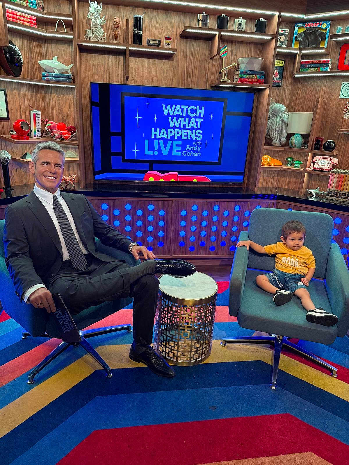 Priyansh is registered to the contest to win money with this photo: man, toddler, child, studio, television, chair, carpet, bookshelf, book, suit, tie, yellow_shirt, denim_shorts, modern, decor, smile, indoors, table, show, colorful