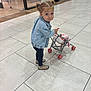 casual_clothing, child, curious_expression, denim_jacket, floor_tiles, footwear, indoor, light, person, pigtails, play, retail_environment, shopping_area, shopping_mall, small_wheels, tile_floor, toddler, toy_stroller, walking, young_child