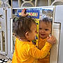 toddler, child, mirror, reflection, yellow_shirt, playpen, indoor, carpet, book, farm_animals, hand, curly_hair, face, smile, toy, plastic, home, baby, play, learning
